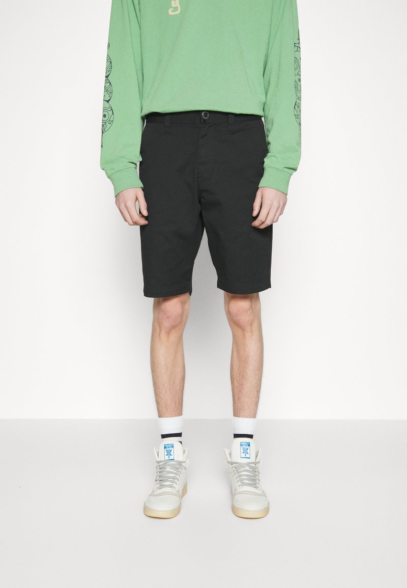 Shorts noirs arrivant au genou avec une coupe décontractée, en tissu mélangé coton. Associés à un maillot à manches longues vert et des baskets blanches montantes.
