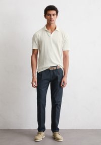 Cream short-sleeve polo shirt, dark trousers, and light beige casual shoes. The shirt has a relaxed collar; the outfit features a brown belt.
