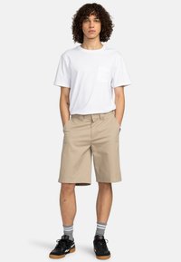 T-shirt bianca in cotone con taschino sul petto, abbinata a pantaloni corti beige fino al ginocchio e scarpe da ginnastica nere, con calzini a righe grigie.