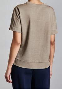 Person som bär en kortärmad, ljusbrun glänsande topp i kombination med mörkblå byxor, sedd bakifrån mot en enkel bakgrund.