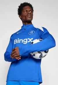 Blå atletisk top med en kvart lynlås, hvide detaljer på ærmerne, og "Bingx" logo. Modellen holder en fodbold og har matchende blå bukser på.
