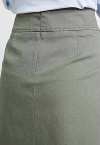 Gonna A-line color khaki realizzata in tessuto testurizzato con vita contenuta e chiusura con zip liscia. Presenta dettagli di cucitura discreti.