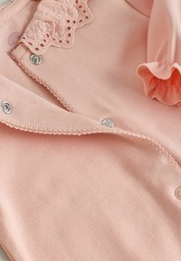 Tela rosa suave con botones a presión plateados, con adorno de encaje festoneado y detalles fruncidos en el borde de la prenda.