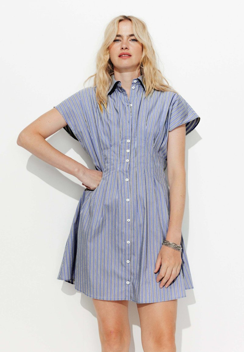 Femme blonde portant une robe chemise à manches courtes bleu et blanche à rayures, avec un col, se tenant avec une main sur la hanche contre un fond blanc.