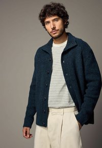 Cardigan din lână de un teal închis cu guler, având butoane mari negre și manșete cu nervuri, așezat peste o cămașă cu dungi crem și pantaloni bej.
