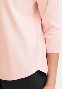 Pull en tricot rose clair avec des manches trois-quarts, des poignets côtelés et une ourlet incurvé, mettant en avant une texture douce et lisse.