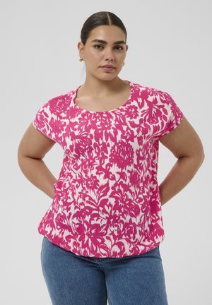 Kobieta o ciemnych włosach, ubrana w różowo-biały kwiatowy top z krótkim rękawem i niebieskie dżinsy, stoi z rękami za plecami na jednolitym tle.