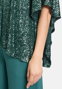 Top verde scintillante in paillettes con design morbido e drappeggiato; presenta un orlo ondulato e maniche trasparenti e aperte. Abbinato a pantaloni teal.
