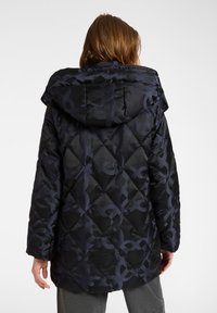 Polstret sort jakke med en blank finish, der har et mørkt mønster, hætte og en firkantet form. Teksturen er glat og quiltet.