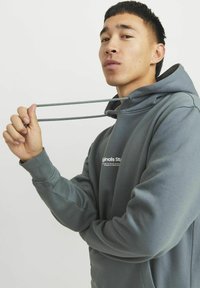 Gray hoodie with a textured finish, featuring a kangaroo pocket and drawstring. Logo printed on the front. Model holds the drawstring.