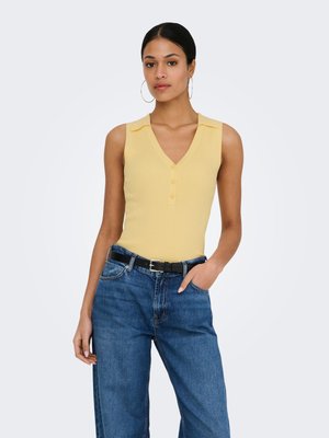 Mujer de pie con la mano en el bolsillo, vistiendo una blusa sin mangas amarilla con cuello y jeans azules con cinturón negro, fondo neutro.