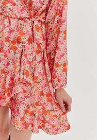 Kvetinové šaty s ružovým základom, zdobené oranžovými a bielymi kvetmi. Mäkka tkanina, úzky pás a detail s volánmi na spodnej časti. Zaviazaná mašľa v páse.