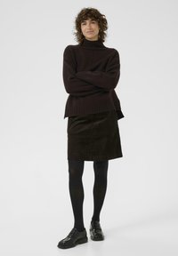 Pull à col roulé tricoté marron foncé, avec un ourlet côtelé, porté avec une jupe en velours côtelé noir. Des collants noirs et des chaussures à enfiler complètent la tenue.