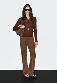 Braun gerippter Langarm-Top, braune Wildlederhosen, dunkelgraue Sonnenbrille und eine kleine braune Umhängetasche. Texturiertes, tailliertes Design mit ausgestellt geschnittenem Bein.