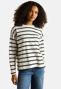 Weißes Langarmhemd mit horizontalen schwarzen Streifen, Rundhalskragen und lockerer Passform, kombiniert mit blauen Jeans.