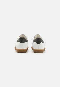 Chaussures en cuir blanc avec un accent noir sur le talon et une semelle en caoutchouc marron clair. Les caractéristiques comprennent une forme arrondie et un logo en relief.