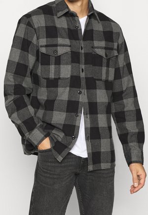 Camisa de franela a cuadros grises y negros con dos bolsillos en el pecho, cierre de botones y mangas largas. Textura suave, corte relajado y patrón clásico.