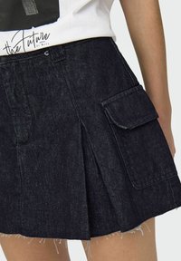 Mörk denimkjol med ett pliserat mönster, utrustad med en framficka och fransad nederkant. Sömnadsdetaljer synliga vid kanterna och fickan.