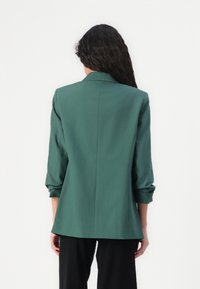 Blazer verde su misura con una silhouette strutturata, maniche tre quarti e una texture liscia, caratterizzato da un risvolto classico e cuciture posteriori.