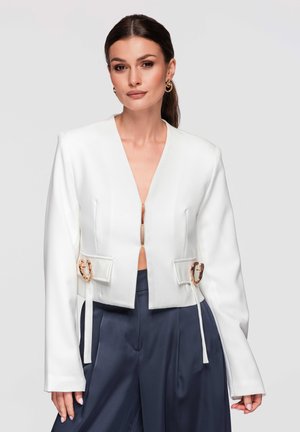 Femme portant un blazer court blanc avec des détails de boucles dorées sur un pantalon large bleu marine, posant devant un fond gris clair.