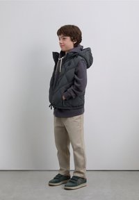 Gray quilted vest with a hood worn over a dark sweatshirt, paired with beige pants and green shoes. Simple design, minimal accents.