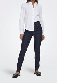 Camisa blanca de botones, jeans ajustados de color azul oscuro con diseño de tres botones, zapatos planos negros con acentos de tachuelas y textura de tela suave.