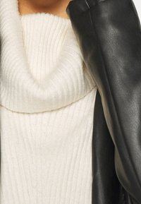 Weißer Rippstrickpullover mit einem Schlauchkragen, darunter geschichtet eine schwarze Lederjacke, die weiche Texturen und kontrastreiche Farben zeigt.