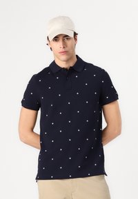 Laivastonsininen polo-paita, jossa valkoinen tähtikuvio, lyhyet hihat ja kaulus. Yhdistetty vaaleanvärisen lippiksen kanssa. Klassinen rento design.