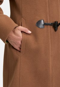 Bruine wollen mixjas met een licht gestructureerde oppervlakte, puntige zwarte leren accenten en zijzakken met een hand erin.