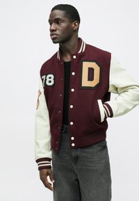 Bordo ir balta universiteto stiliaus striukė su „D“ emblema, numeriu „78“, spaudžiamomis sagomis, dryžuotu apykakliu ir tekstūruotomis rankovėmis.