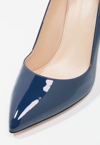 Escarpins stilettos en cuir verni bleu marine avec un bout pointu et une surface lisse, présentant un léger éclat et une doublure intérieure de couleur claire.