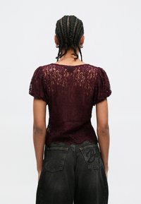 Top de renda borgonha com mangas curtas bufantes, padrão floral texturizado e bainha com acabamento em ondas. Combinado com calças de ganga escuras com bordados decorativos.