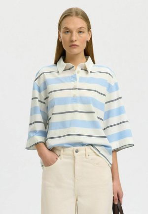 Žena s rovnými vlasmi nosí polo tričko so širokými rukávmi s modrými, bielymi a čiernymi vodorovnými pruhmi, béžové nohavice, drží tmavohnedú tašku.