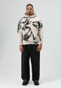 Beige tie-dye hoodie met een kangoeroezak en logo, gecombineerd met loszittende zwarte jeans en zwarte veterschoenen.