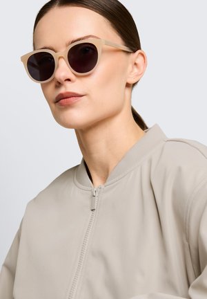 Femme aux cheveux plaqués en arrière portant une veste bomber beige et des lunettes de soleil foncées à monture beige sur fond clair.