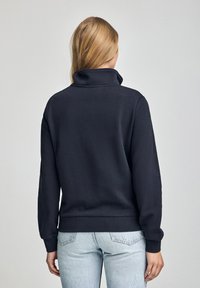 Svart fleece-tröja med hög krage, ribbade ärmar och nederkant, buren med ljusblå denimjeans som visar en slät textur.