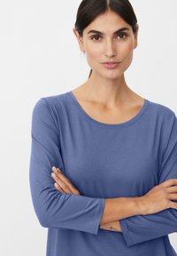 Niebieska bluzka z długim rękawem, okrągłym dekoltem i teksturowaną tkaniną. Prosty design, noszona przez osobę ze skrzyżowanymi ramionami.