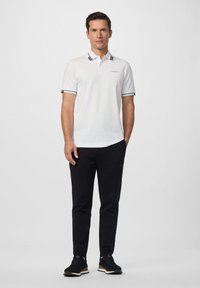 Muž stojící v bílém polo tričku s krátkým rukávem a černých kalhotách, obutý v černých teniskách, na jednobarevném světle šedém pozadí.