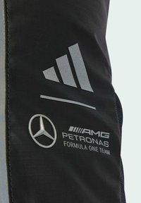 Čierna látka s strieborným logom Adidas s tromi pruhmi, emblémom Mercedes-Benz a textom AMG Petronas Formula One Team, s textúrovaným povrchom.