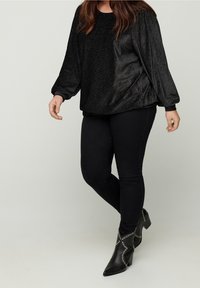 Kobieta ubrana w czarny, błyszczący, długi rękaw top, czarne skinny spodnie i czarne, szpiczaste botki za kostkę z srebrnymi ćwiekami, stojąca na jednolitym tle.