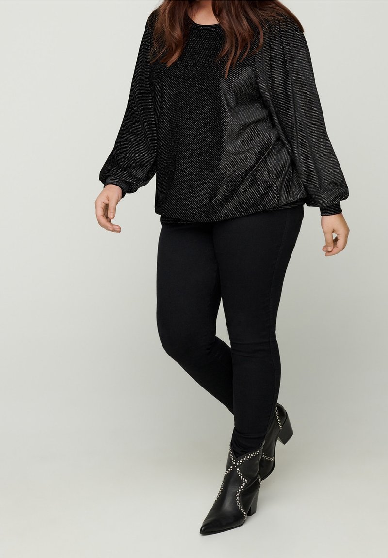 Donna che indossa un top lungo a maniche nere luccicanti, pantaloni skinny neri e stivaletti neri con punta e borchie argento, in piedi contro uno sfondo semplice.