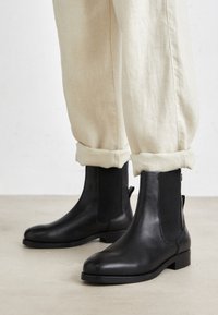 Bottines en cuir noir avec des panneaux élastiques sur les côtés, bout arrondi, talons bas en bloc, portées avec un pantalon beige retroussé.