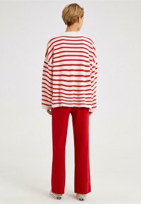 Person mit kurzen Haaren, der einen weiß-rot gestreiften Pullover, rote Hosen und weiße Turnschuhe trägt und vor einem schlichten Hintergrund steht.