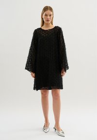 Schwarzes, strukturiertes Kleid mit langen Ärmeln, rundem Halsausschnitt und unregelmäßigem Saum. Kombiniert mit silbernen, spitzen High Heels.