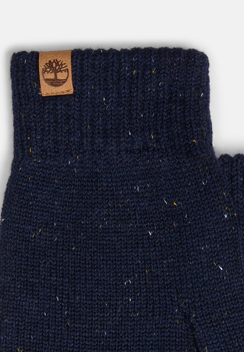 Guantes de lana en color navy con un puño acanalado y motas amarillas y blancas dispersas. Presentan un parche de logo de cuero marrón en la zona de la muñeca.