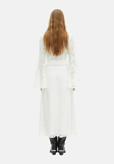 Vrouw met lang haar in een witte kanten jurk met uitlopende mouwen en zwarte enkellaarsjes, staand met de rug naar de camera tegen een effen achtergrond.