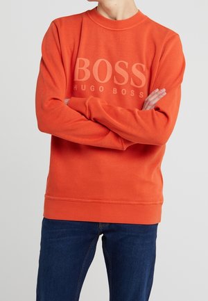 Person iført en orange Hugo Boss sweatshirt med armene over kors og blå jeans, vist fra nakken til midt på låret mod en ensfarvet baggrund.