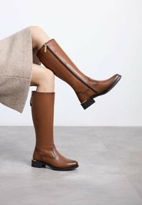 Botas de cuero marrón hasta la rodilla con cremallera lateral, tacón bajo y detalles de costura en la parte delantera y trasera. Forro interior texturizado visible.