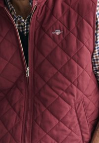Burgundy gewatteerde vest met een rits aan de voorkant en zijzakken, met een klein logo op de borst en een geblokt shirt dat eronder zichtbaar is.