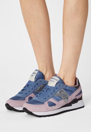 Pieds chaussés de baskets Saucony Shadow bleues et roses avec des semelles noires et blanches, présentés sur un fond clair uni.